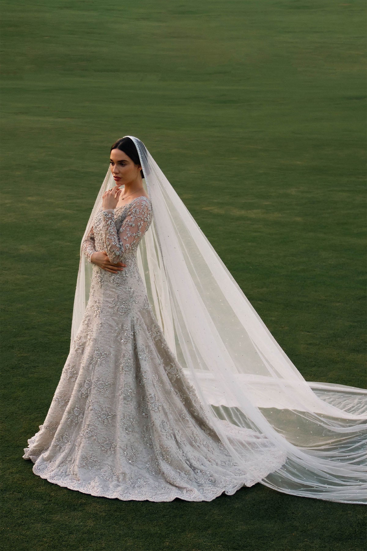 Model in a side shot wearing a net bridal gown with a mermaid fit and train and a cathedral-style veil with a pearl spray