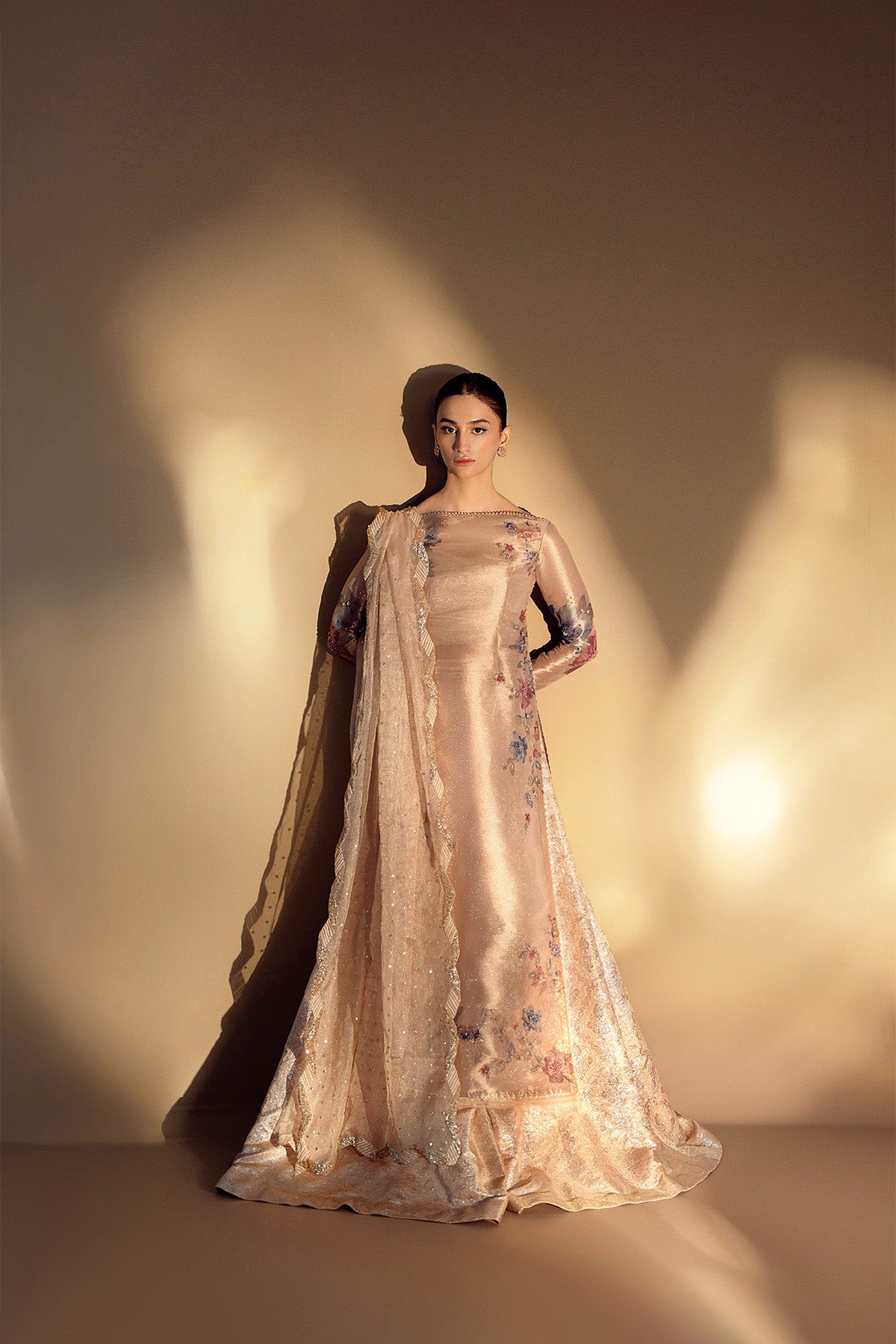 A woman wearing a beige and gold traditional outfit with floral prints, zardozi work, and lace trimming, consisting of a shirt, sharara pants, and a dupatta with a scalloped border.