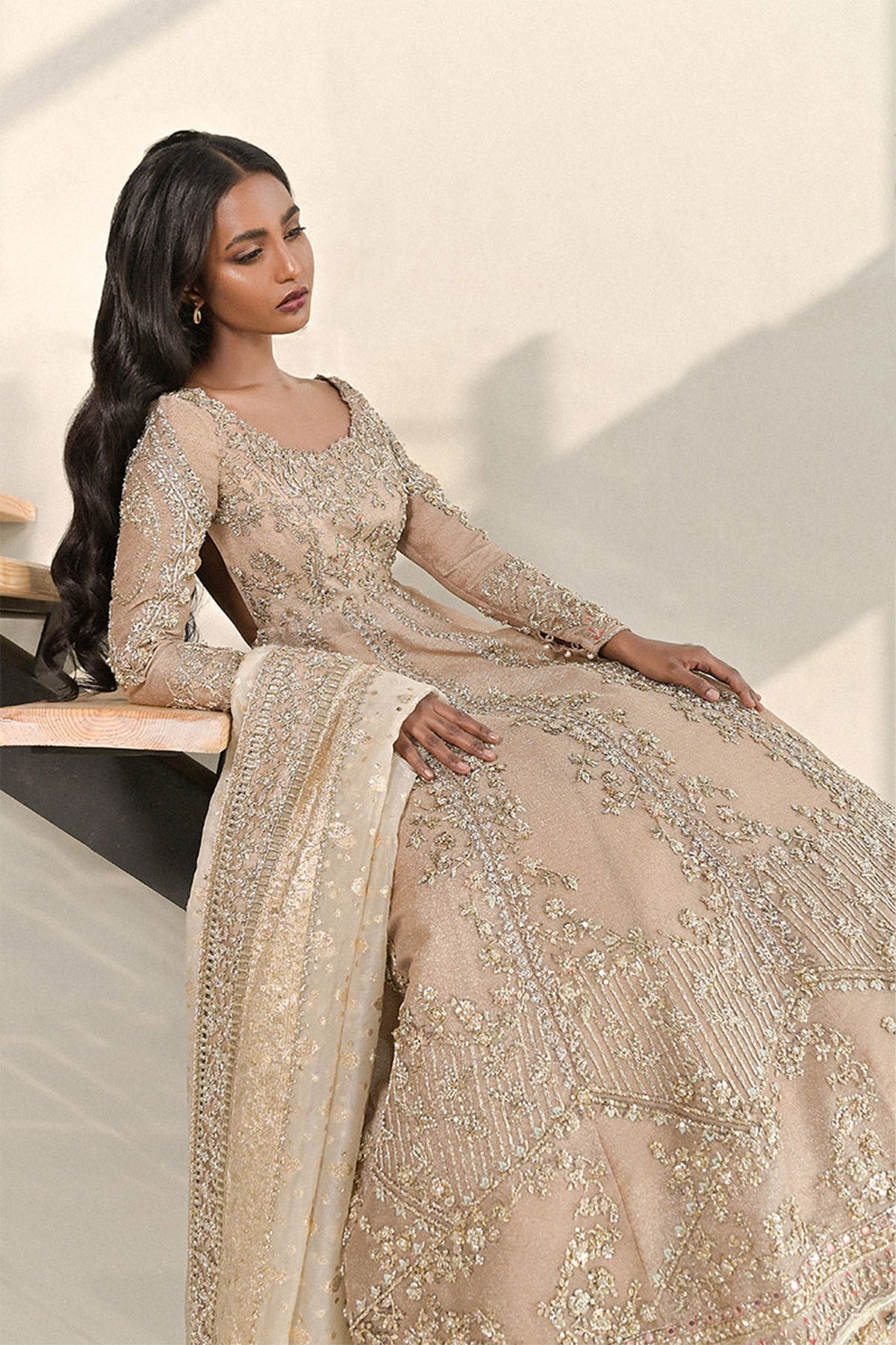 Model in a seated side shot wearing a dusty pink pishwas paired with lehenga along with an organza zari dupatta with colored and embellished borders