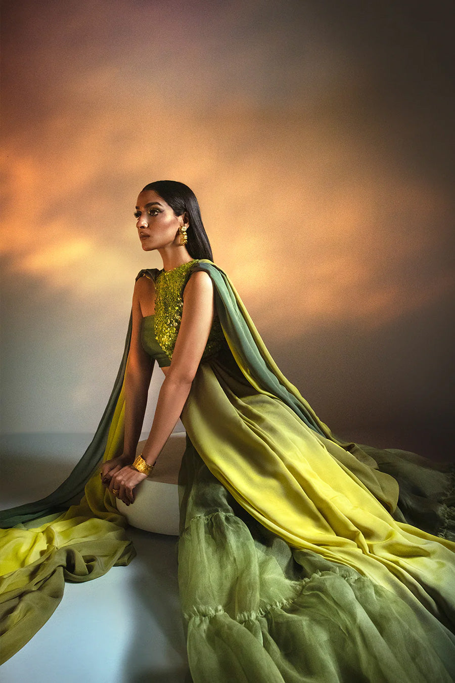 Model in a seated side view shot wearing a lime green halter blouse paired with an ombre drape in Italian chiffon silk and a ruffled silk organza skirt