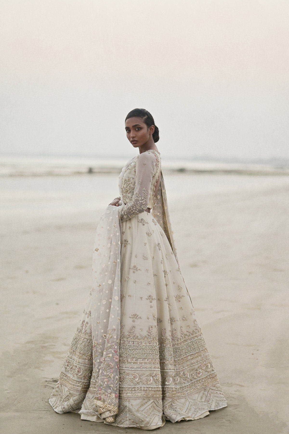 Model in a side view shot wearing long pishwas gown in organza silk paired with an organza zari dupatta and organza lehenga