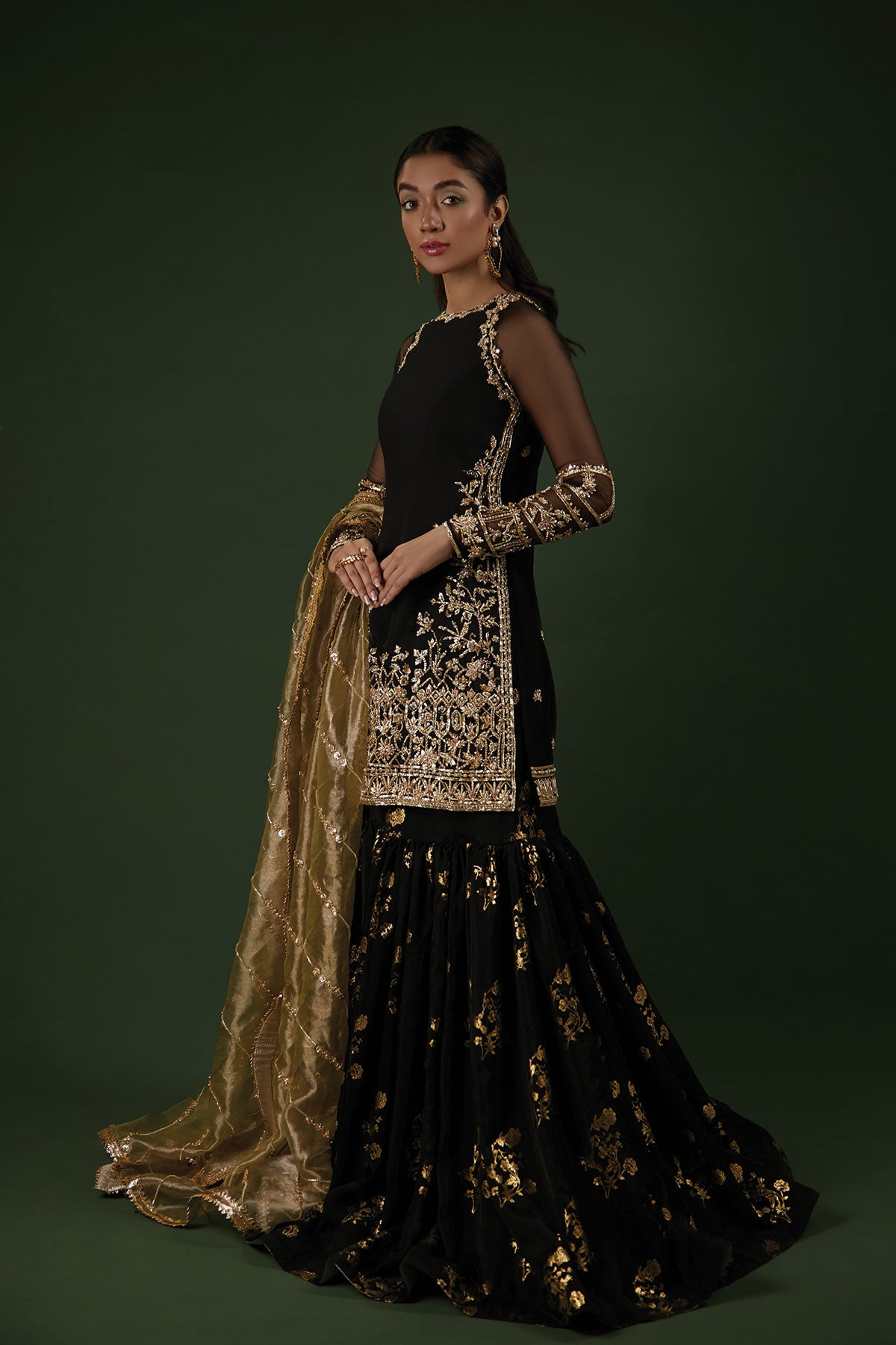 Model in a back shot wearing a black organza shirt paired with a silk chiffon foil printed gharara and an olive green tissue dupatta