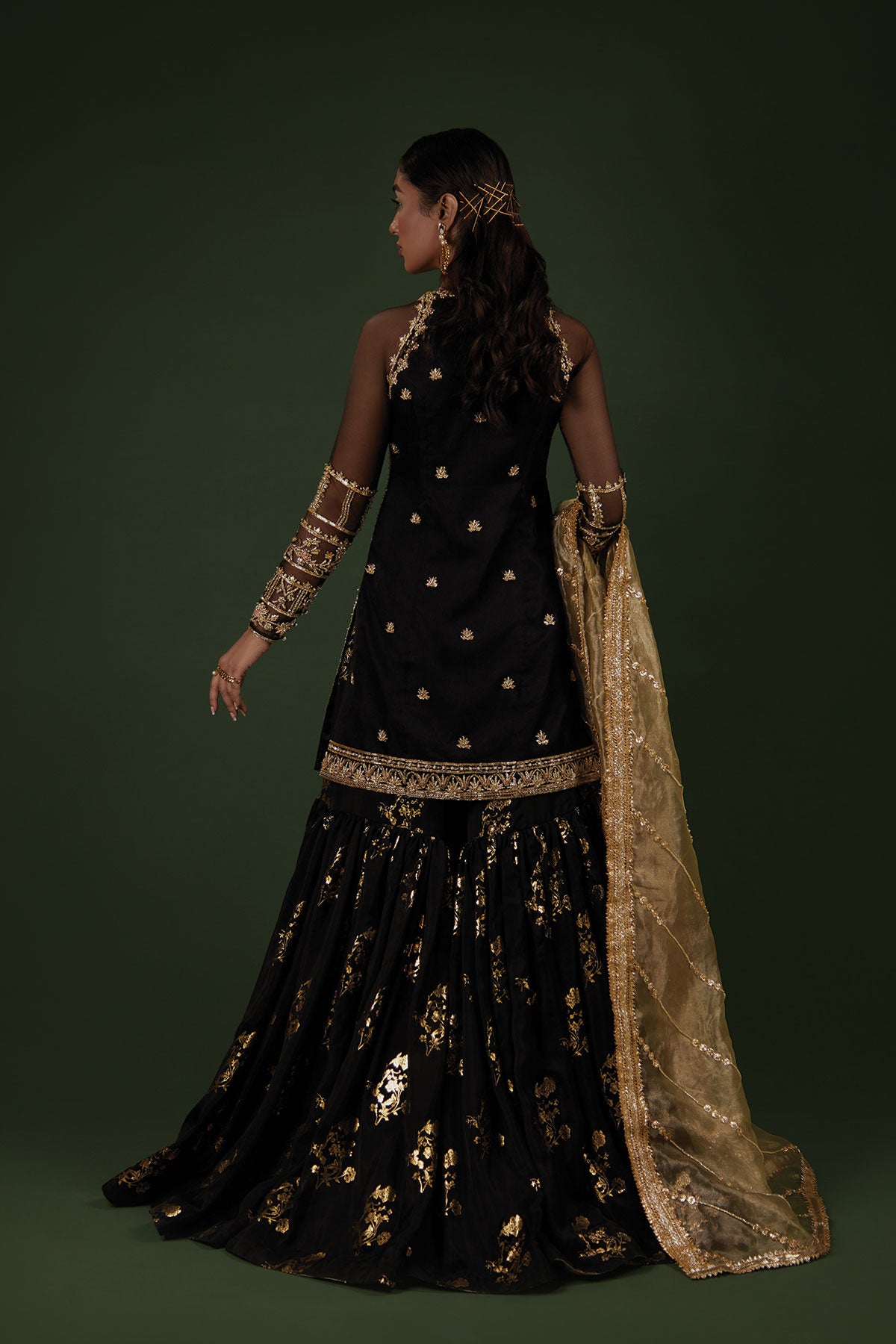 Model in a side shot wearing a black organza shirt paired with a silk chiffon foil printed gharara and an olive green tissue dupatta