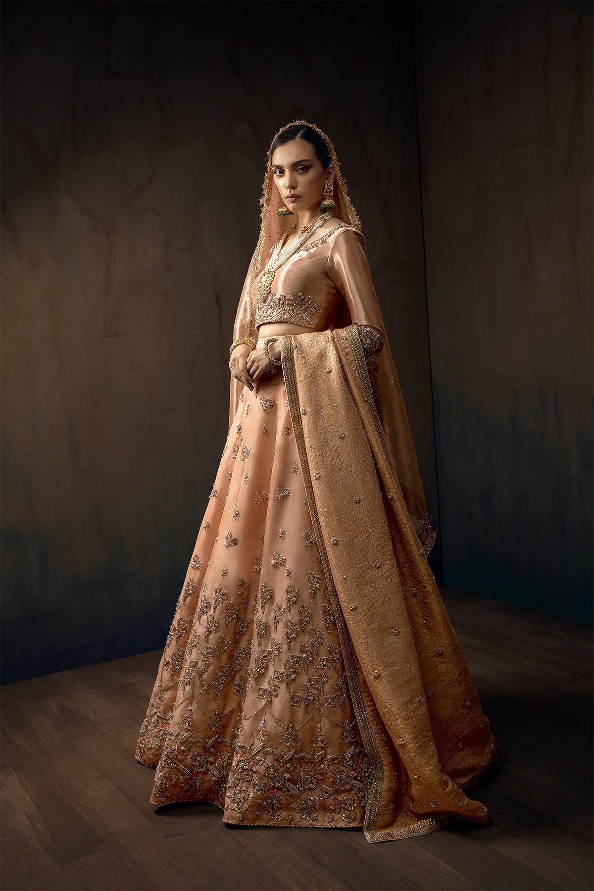Model in a full side shot wearing melon pink lehenga in organza fabric paired with a pure tissue choli, dupatta accompanied by a jamavar shawl