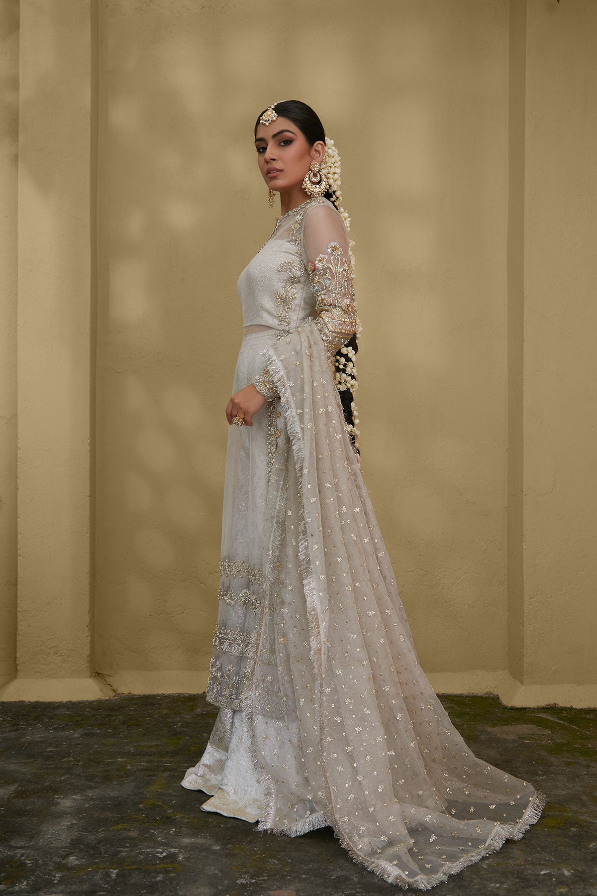 Model in a side shot wearing white kundan net shirt paired with silk brocade sharara pants and a bustier and a kundan net dupatta