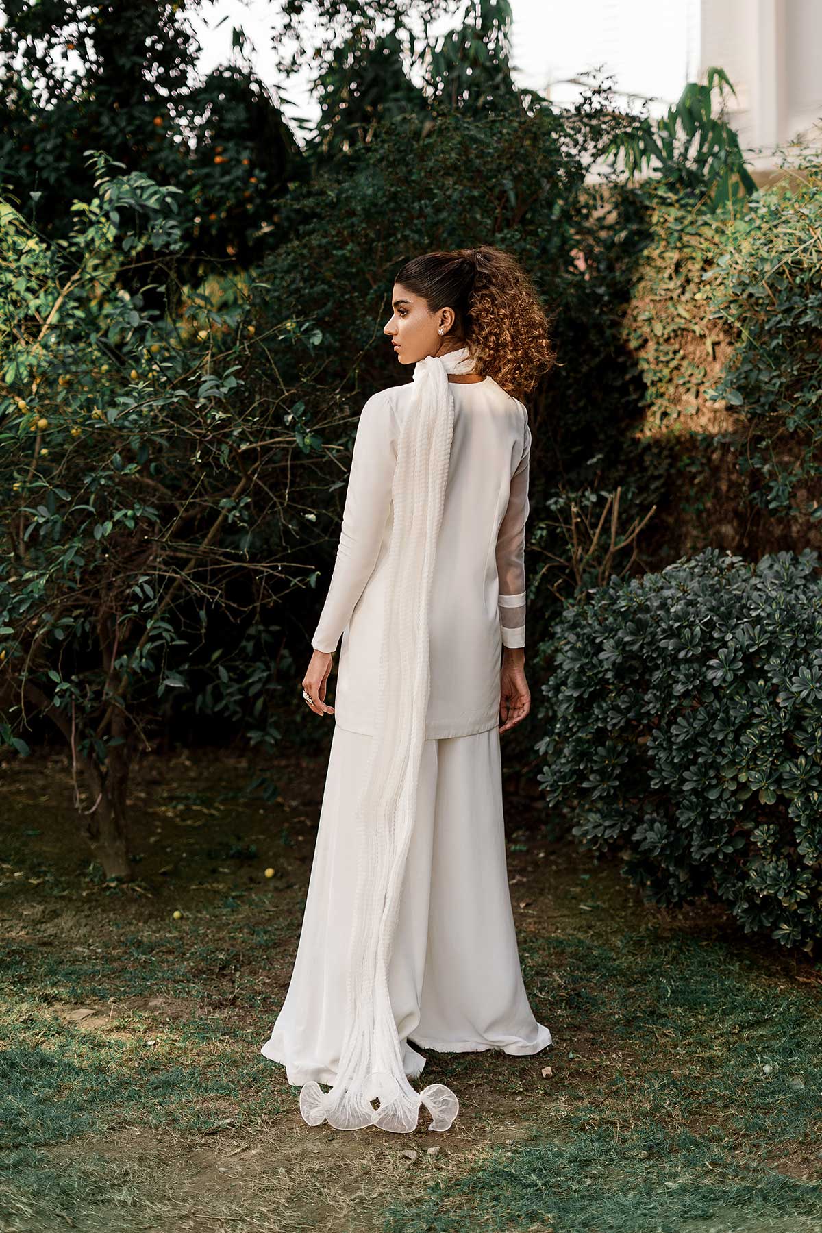 "Model in a back shot waering jacket with a sheer organza side paired paired with flared pants, a bustier, and a crushed dupatta