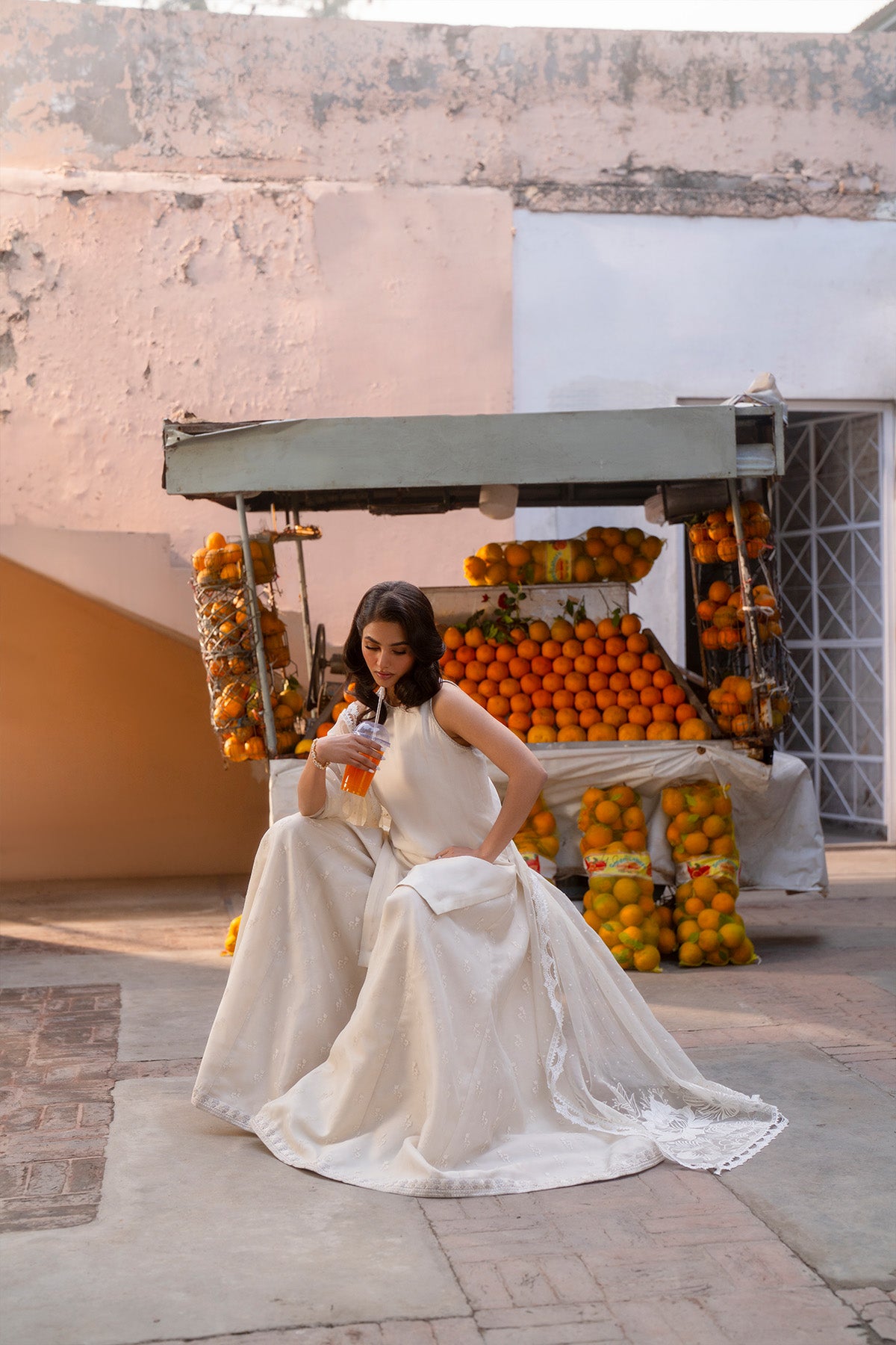 Model in a front shot sitting on a bench wearing a beige silk shirt paired with screen-printed organza sharara pants.