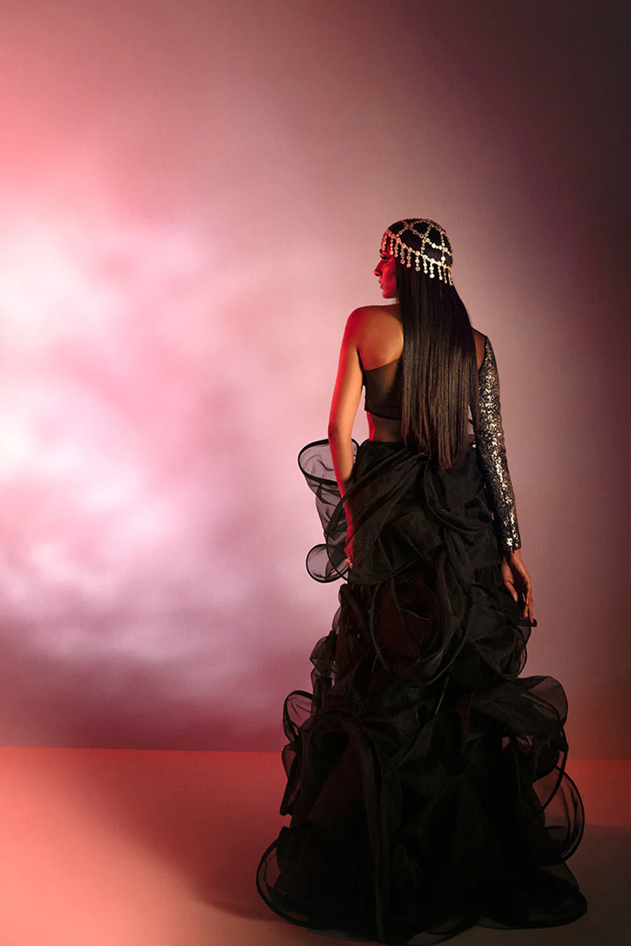 Model in a back shot wearing a black formal silk blouse with silver sequins and a sheer black back, paired with a fully ruffled black organza skirt