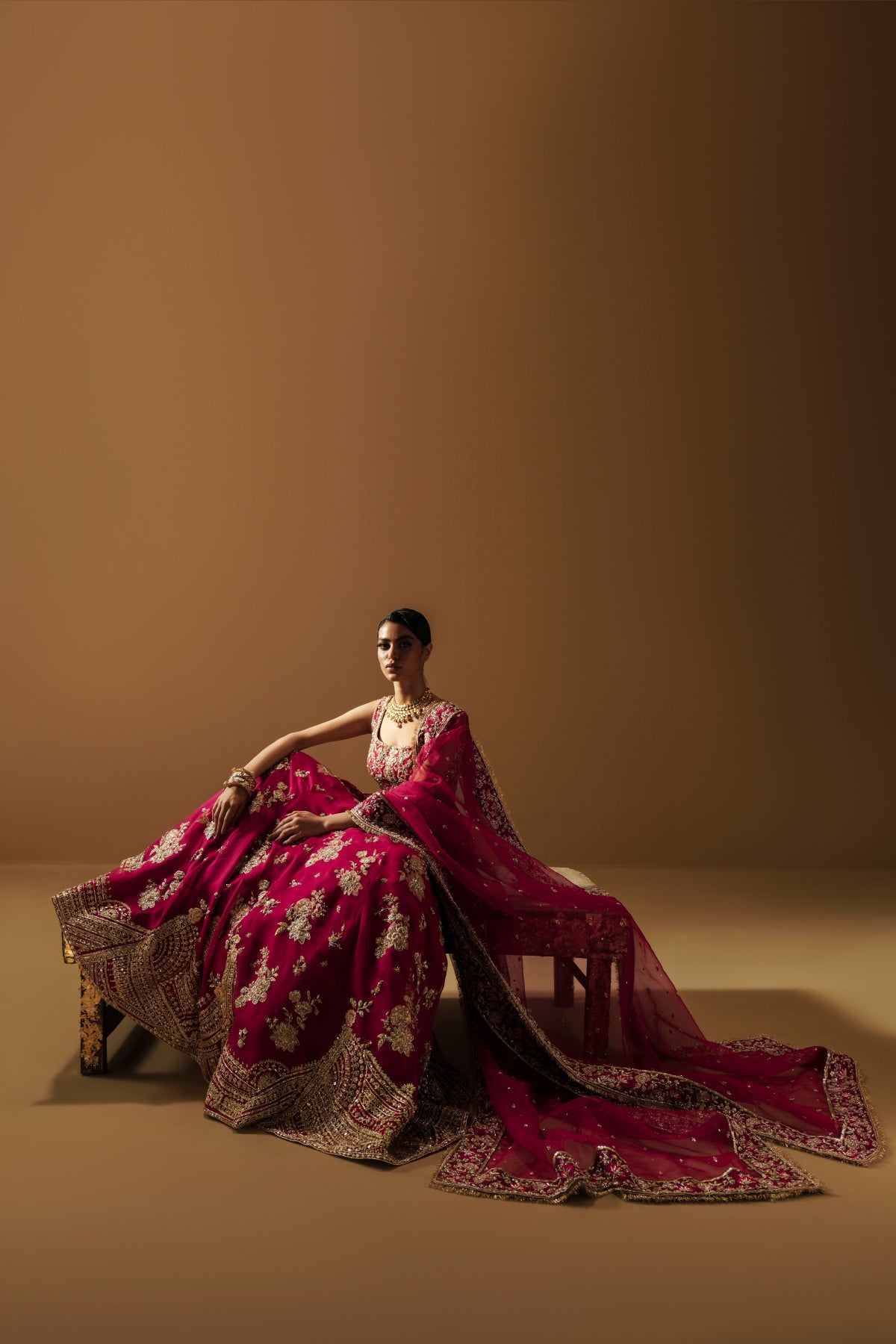 Model in a seated front view angle wearing a dark pink lehenga choli with handcrafted zardozi and sequin work and an organza dupatta