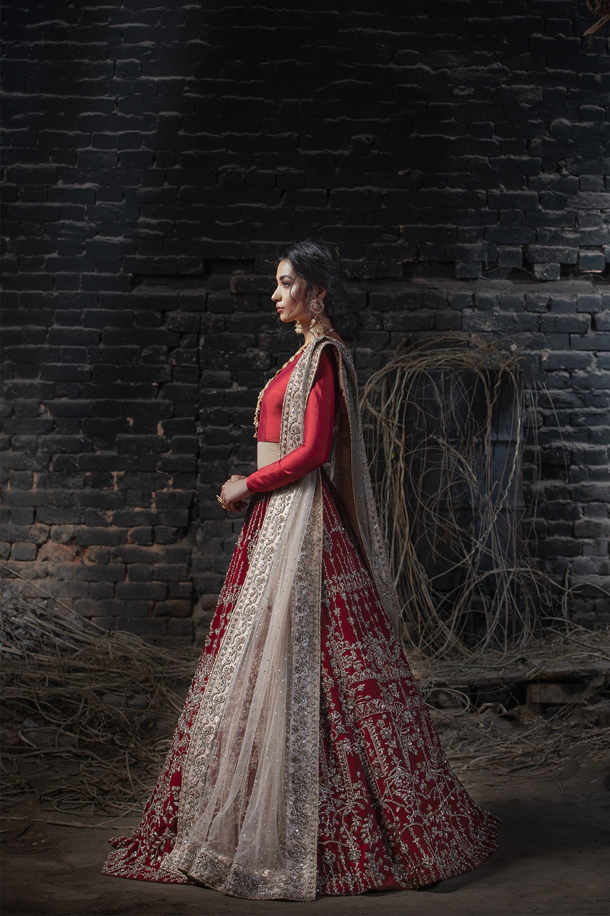 Model in a side view shot wearing deep red velvet lehenga paired with red silk choli along with a nude pink dupatta with heavily embellished borders