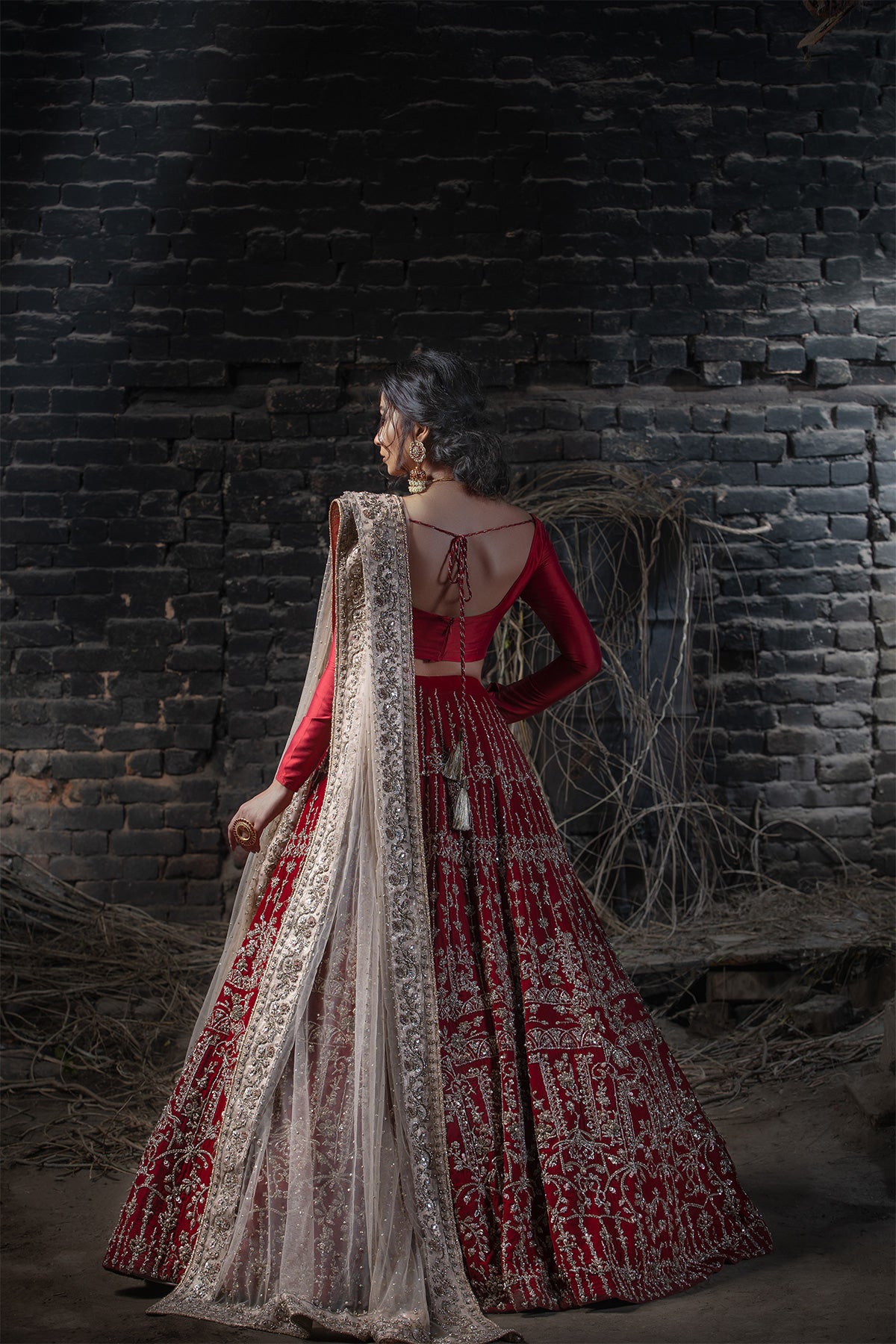 Model in a back shot wearing deep red velvet lehenga paired with red silk choli along with a nude pink dupatta with heavily embellished borders