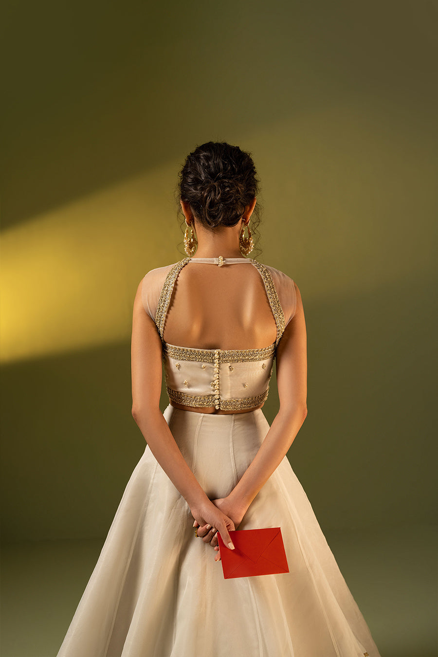 Model in back shot wearing ivory choli paired with organza lehenga holding a red envelope 