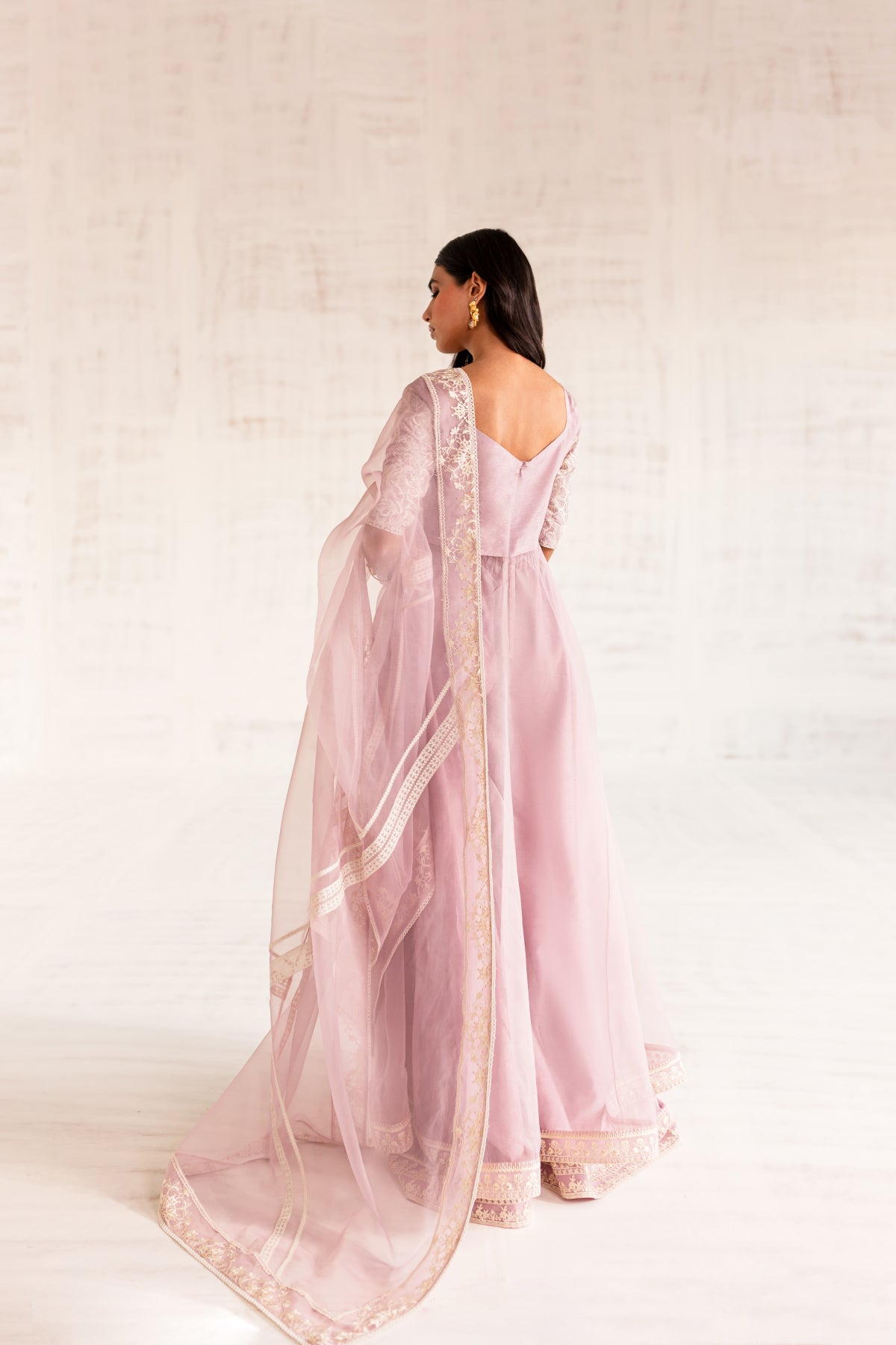 Model in a back view shot wearing an organza light pink long flared pishwas paired with an organza dupatta and silk katan trousers