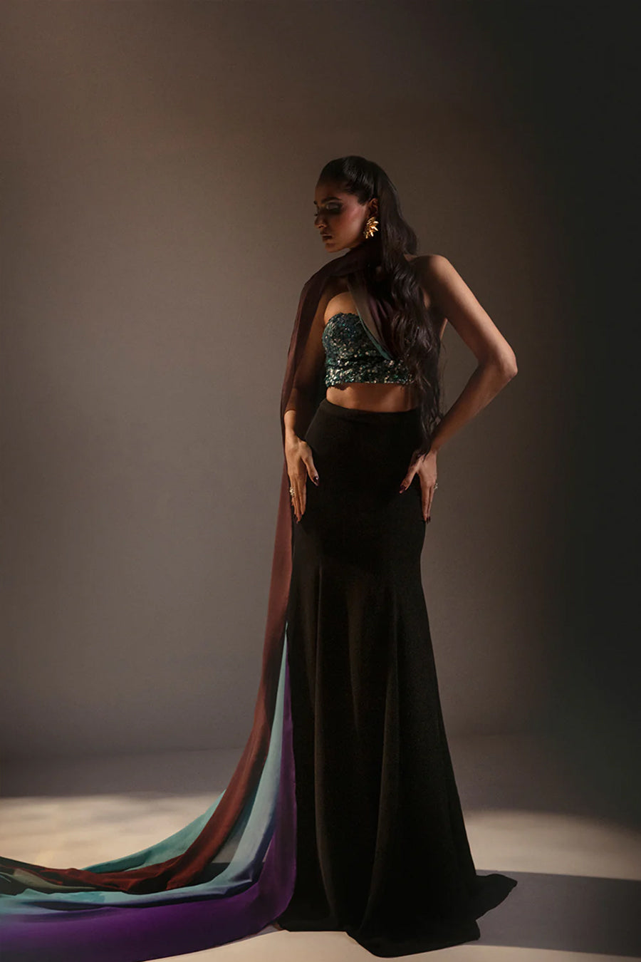 Model in a front shot of a multi-colored blue corset blouse embellished with sequins, paired with a fitted black skirt and a silk chiffon ombre drape