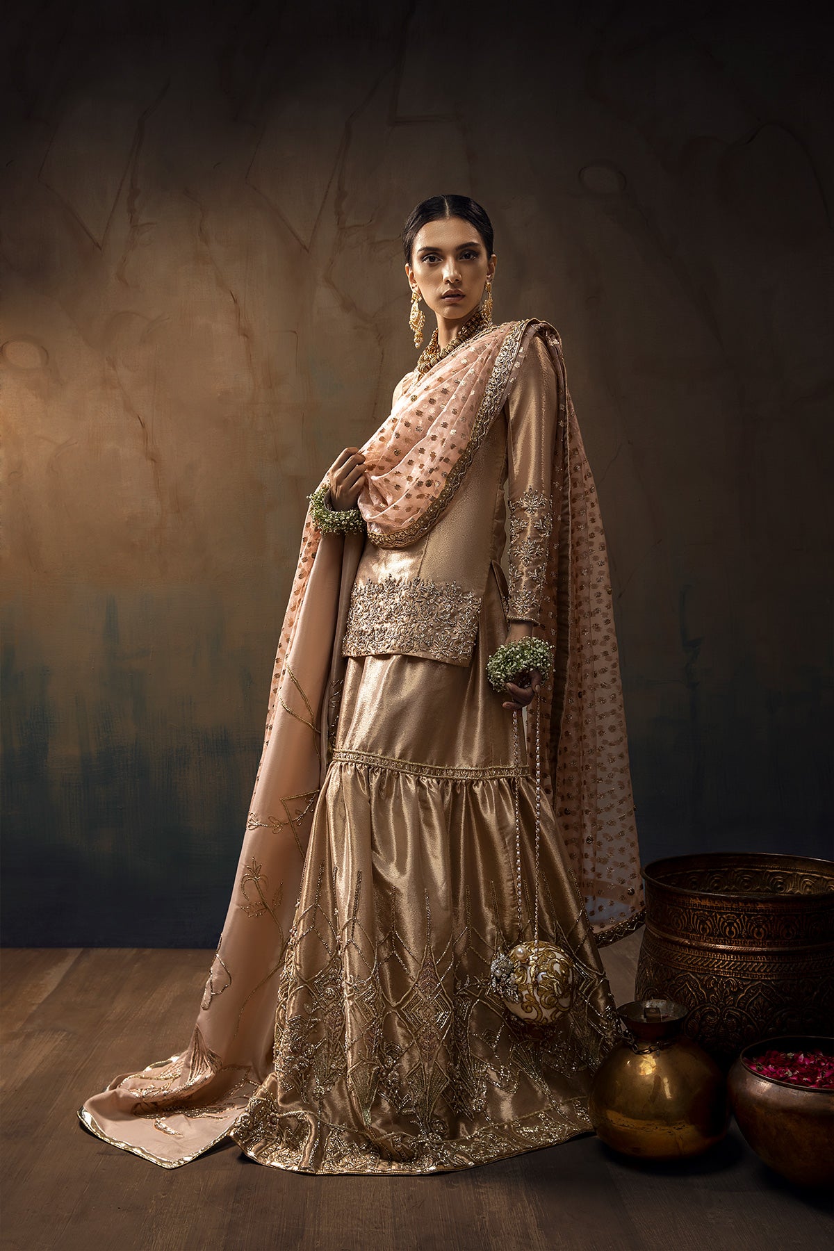 Model in a side full shot wearing a rose gold bridal gharara with rose gold shirt paired with blush pink silk organza zari dupatta and a silk shimmer shawl