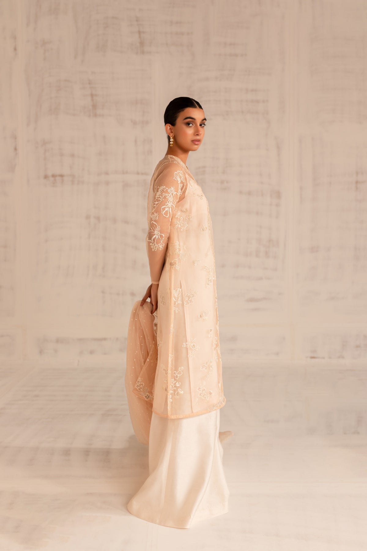 Model in a side view shot wearing a warm coral organza jacket-styled shirt paired with an organza dupatta and silk katan pants