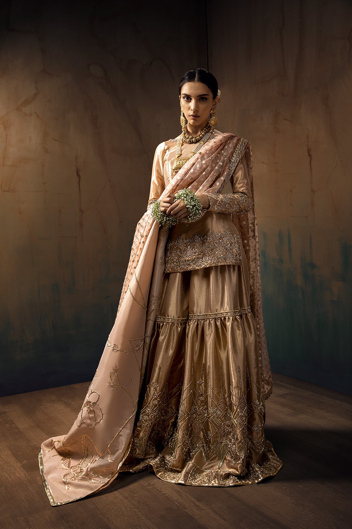 Model in a front full shot wearing a rose gold bridal gharara with rose gold shirt paired with blush pink silk organza zari dupatta and a silk shimmer shawl