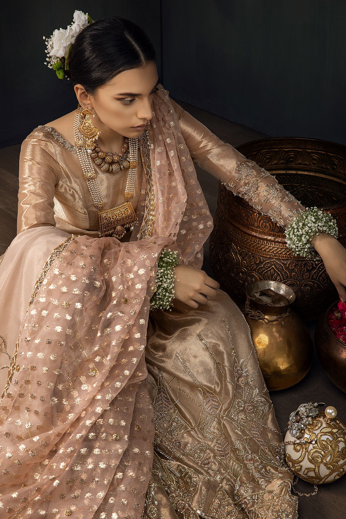 Model in a seated top shot wearing a rose gold bridal gharara with rose gold shirt paired with blush pink silk organza zari dupatta and a silk shimmer shawl