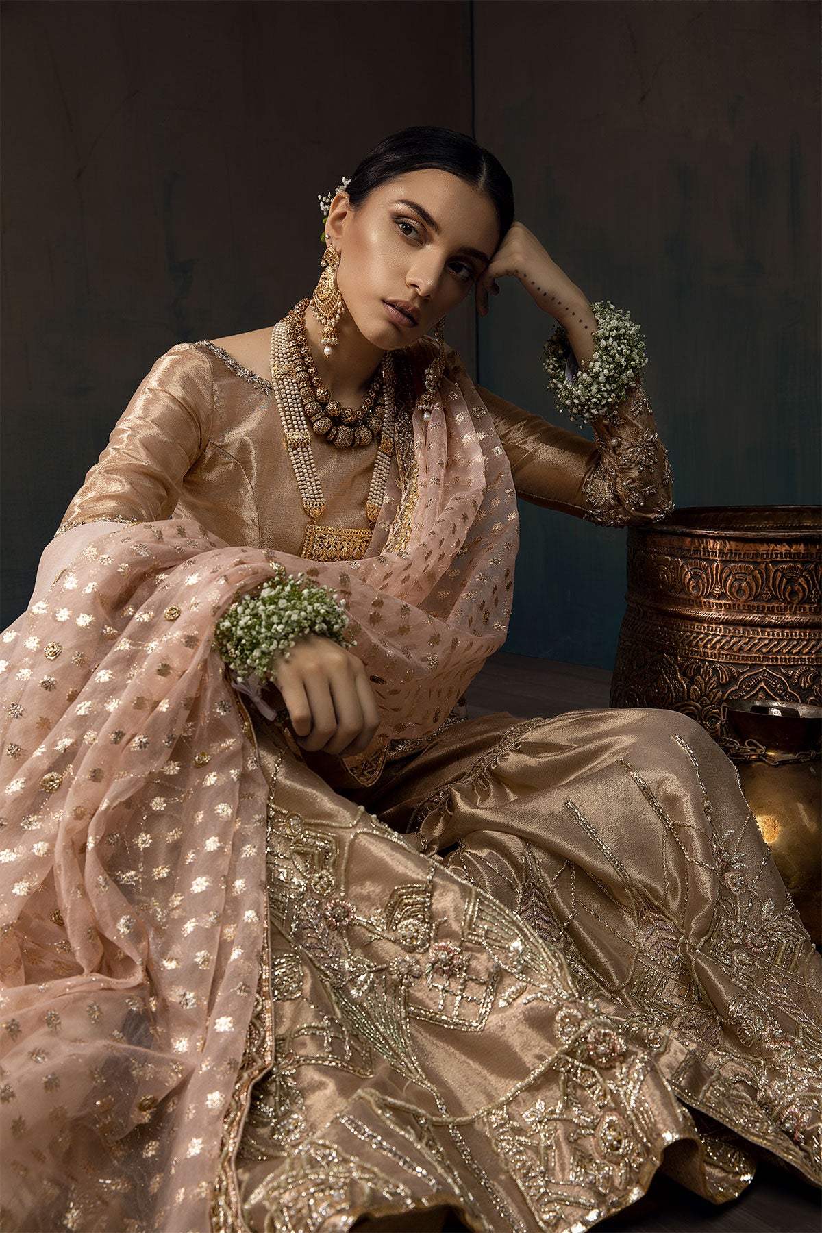 Model in a seated shot wearing a rose gold bridal gharara with rose gold shirt paired with blush pink silk organza zari dupatta and a silk shimmer shawl