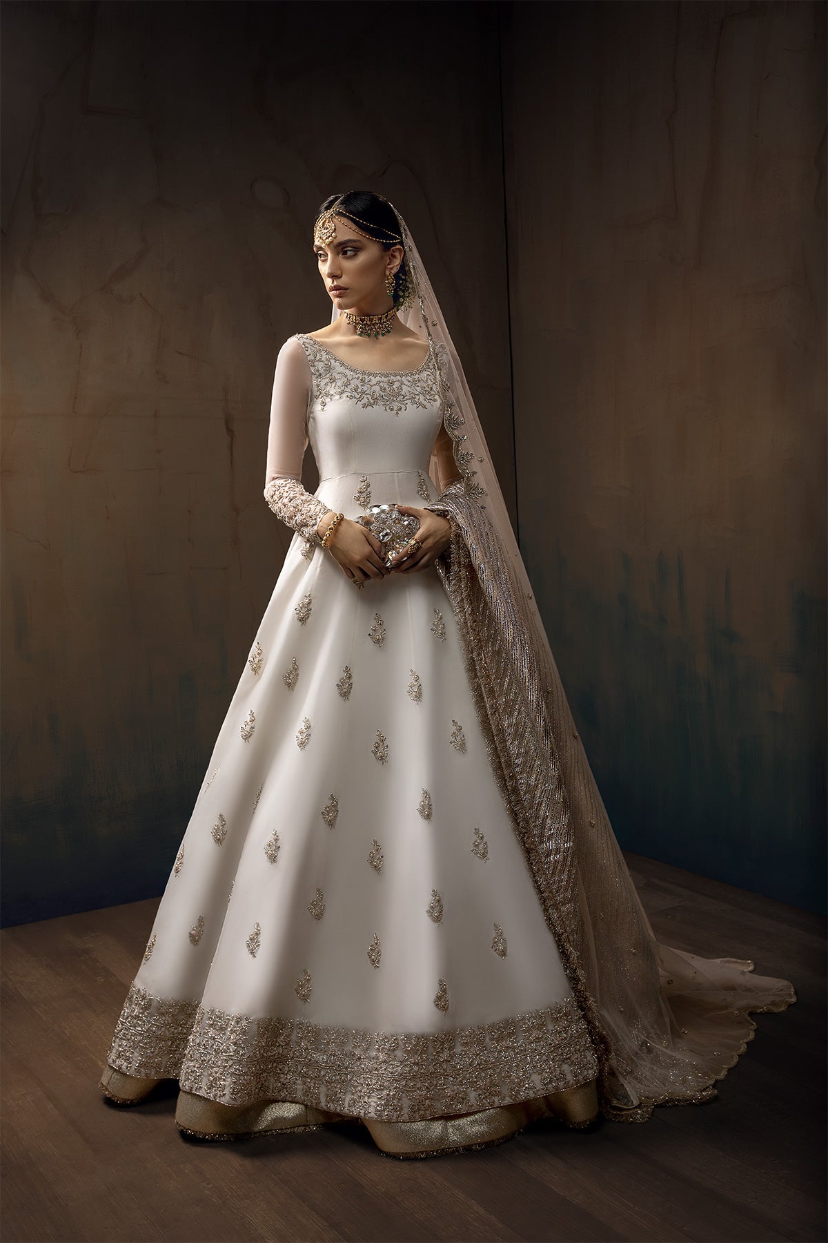 Model in a front full shot wearing white long pishwas gown paired with lehenga and net dupatta along with a silver shawl 