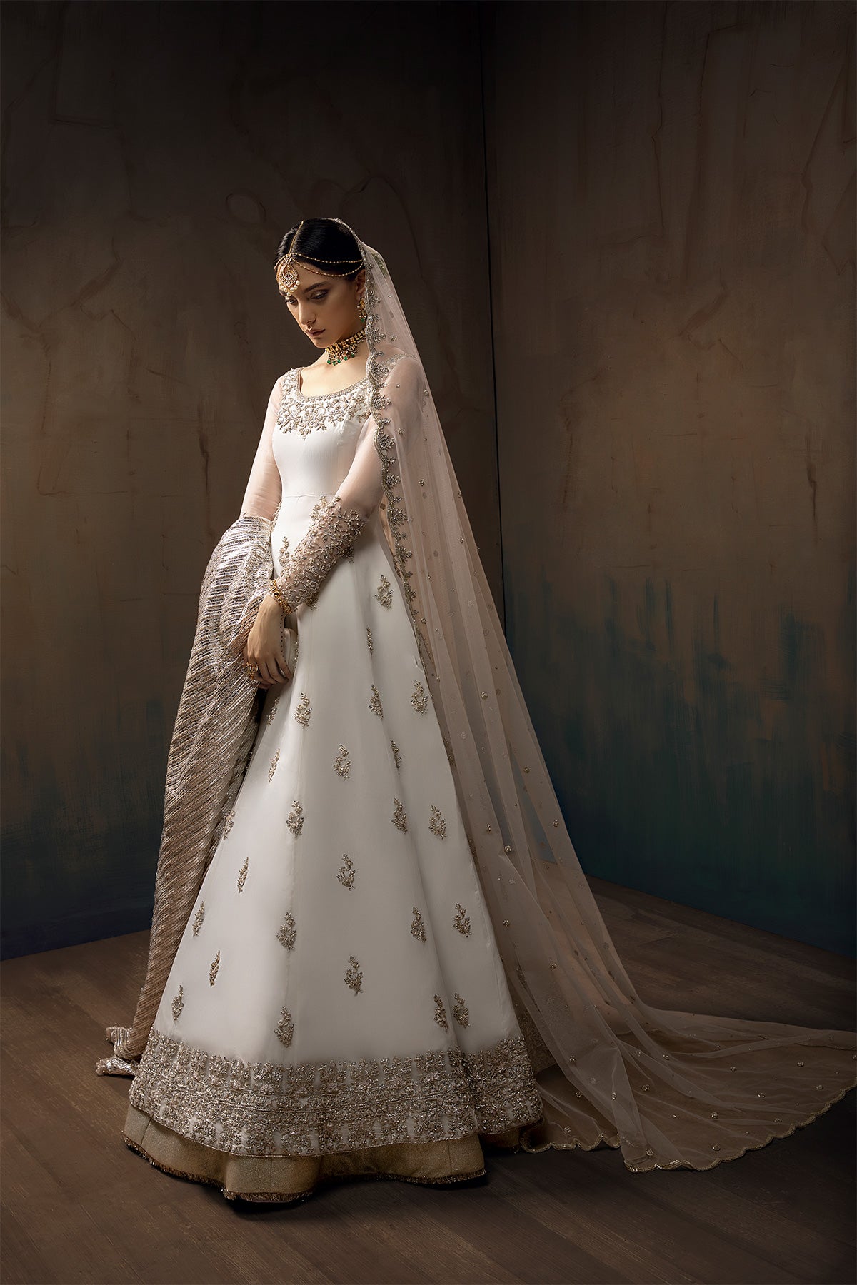 Model in a front full side shot wearing white long pishwas gown paired with lehenga and net dupatta along with a silver shawl 